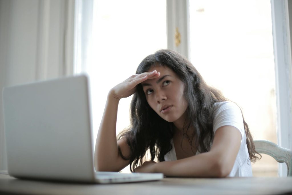 Irritated ethnic female entrepreneur in casual wear sitting at table with netbook and touching head while waiting for internet connection during remote work